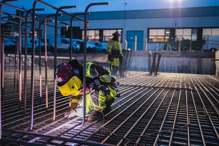 Welding workers at construction site, reinforcing steel rebar installation for concrete foundation, safety gear on, urban commercial building in background, early morning or evening lighting, construction project.